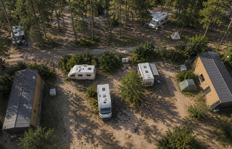 Luftaufnahme eines Campingplatzes im Wald mit Wohnmobilen, Tiny Häusern und schattigen Stellplätzen.